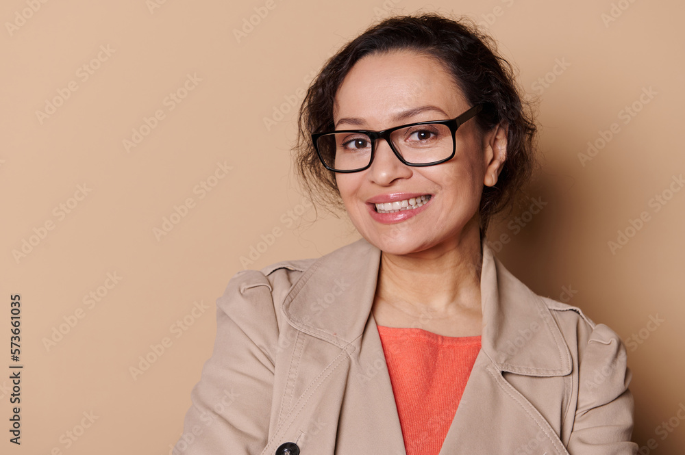 Closeup portrait of a middle-aged ethnic pretty woman in beige coat, wearing stylish eyeglasses, smiling with a beautiful a toothy smile looking at camera, isolated on cream background. Ophthalmology Closeup portrait of a middle-aged ethnic pretty woman in beige coat, wearing stylish eyeglasses, smiling with a beautiful a toothy smile looking at camera, isolated on cream background. Ophthalmology