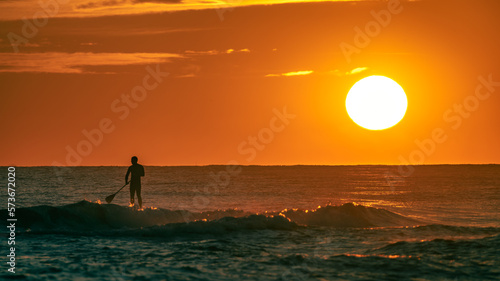 Silhouette of a man practicing paddle surfing on a wave in the sea with the sunset and the sun sphere in the background. Castelldefels  Barcelona