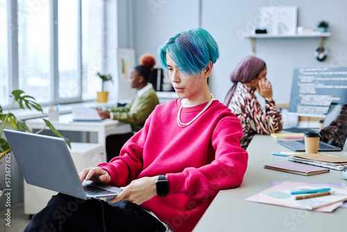 Portrait of Asian young man with colored hair using laptop in modern office, magenta accent photo