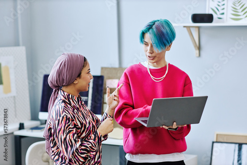 Waist up portrait of creative gen Z team discussing project while standing in office and holding laptop