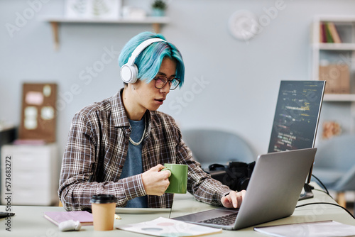Fotografiet Portrait of young man with blue hair using computer in office and wearing headph