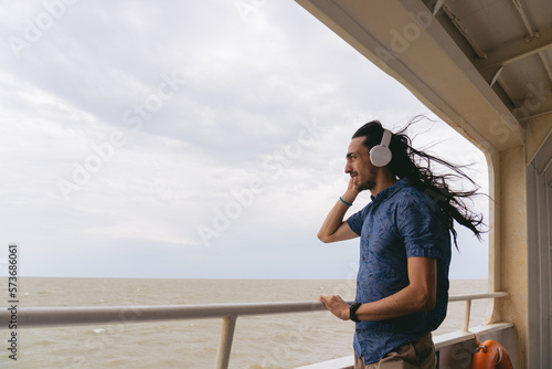 Young Latin man with long hair listening music with headphones on the deck of a ferry. Copy space.