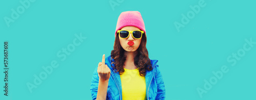 Portrait of bad girl expression showing hand with middle finger sign wearing colorful clothes, pink hat, sunglasses on blue background