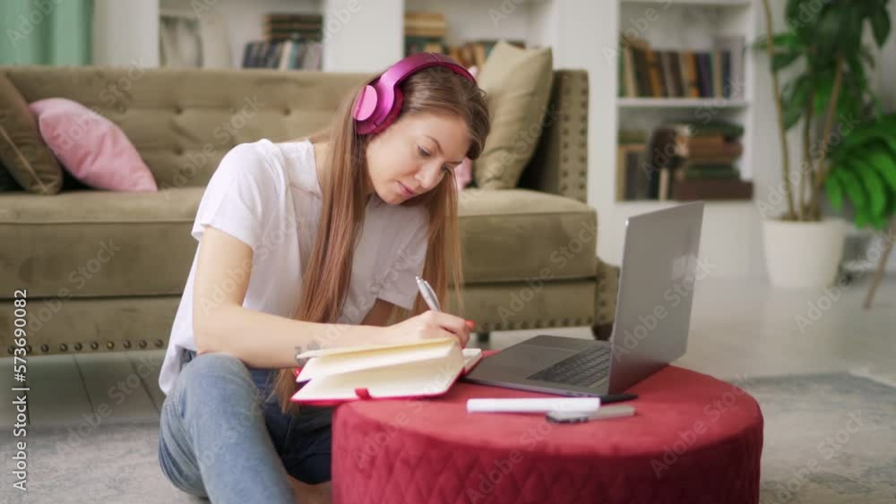 Girl student studying at home reading notes in exercise book using computer for online self education doing homework. Young woman with blond hair in headphones reading a book and taking notes. 4k