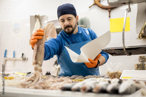 Fotografía middle-aged fishmonger in apron and hat is hand picking a squid from the counter to sell it to a customer, selling food, small and medium business