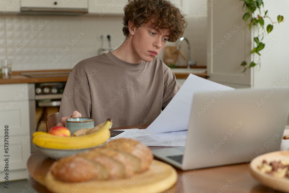 Portrait of a young curly haired casual-dressed male student having a home-cooked breakfast sitting at a table focused with papers preparing for exam using laptop in morning in light kitchen indoors.
