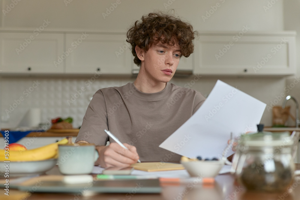 Close up of young curly haired handsome man wearing beige t-shirt sitting at a kitchen table surrounded by comforts of domestic life making notes reviewing papers while having breakfast in kitchen. Close up of young curly haired handsome man wearing beige t-shirt sitting at a kitchen table surrounded by comforts of domestic life making notes reviewing papers while having breakfast in kitchen.
