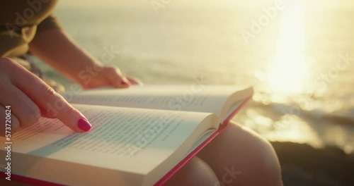 Girl self-learning by reading a book outdoors. Ocean waves on the beach at sunset. Close-up of woman hands. Finger points to printed text. photo