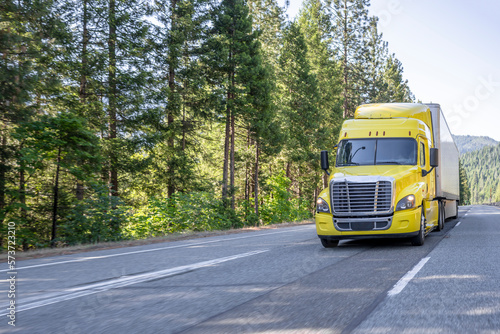 Industrial standard yellow big rig bonnet semi truck transporting goods in dry van semi trailer driving on the interstate highway road