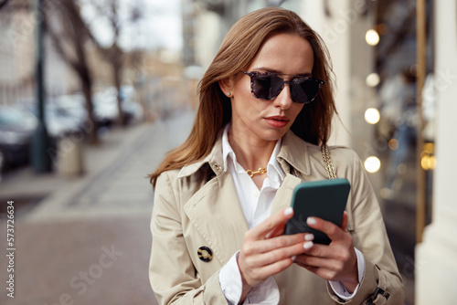 Fashionable woman in sunglasses is standing with phone on city street background 