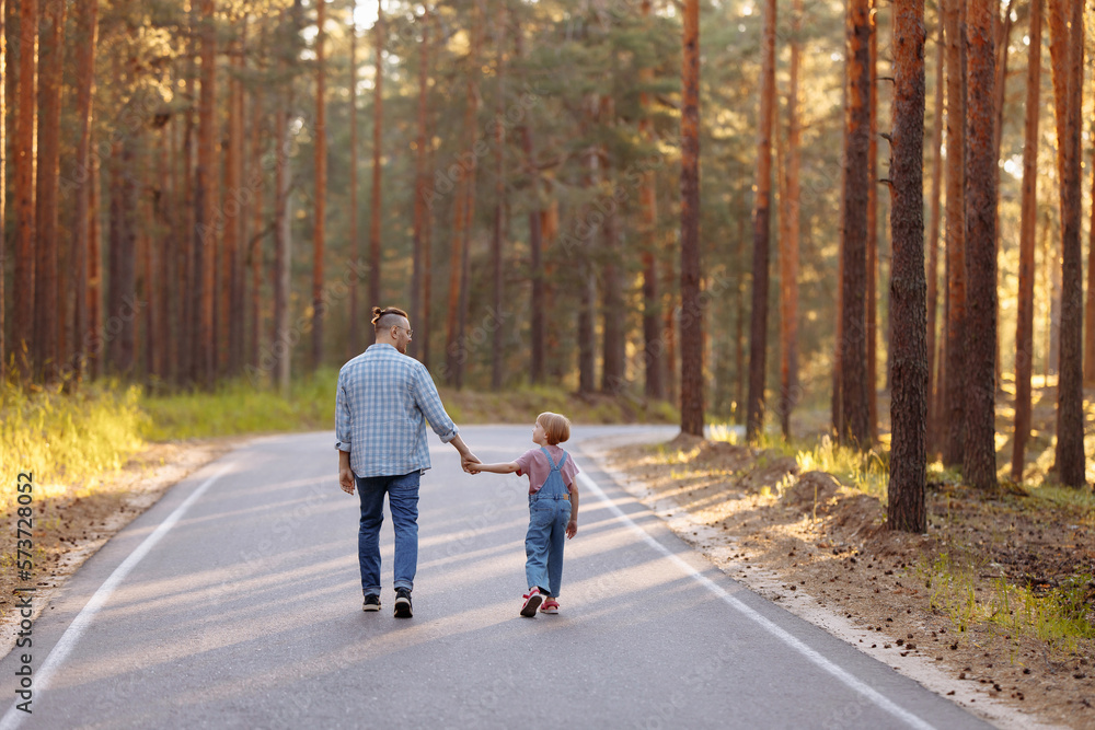 Fototapeta premium Dad and his little daughter are walking along a forest road among tall pines. Family walk in the forest at sunset, man and little girl.