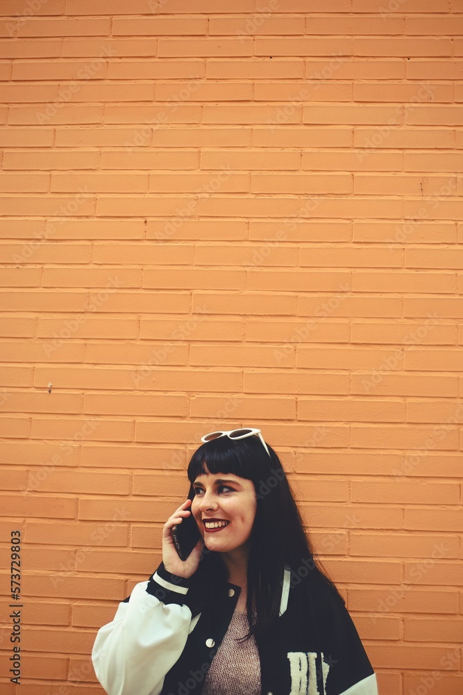 Naklejka premium Young millennial girl smiling while talking on cellphone with friend looking to the side. Copyspace on top of image.