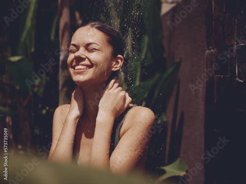 Fotografiet A woman, a body in a swimsuit washes her head in a tropical shower outdoors against backdrop green tropical leaves, flowers and palm trees