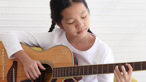 Cute happy Asian girl with long hair braided in pigtails playing acoustic guitar and singing a song in living room, enjoy spend time on music hobby, learn and practice playing guitar instrument