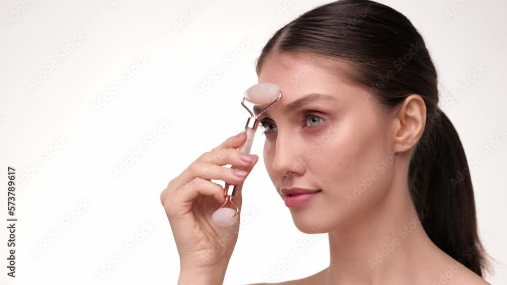 Close up portrait healthy young caucasian woman using jade facial roller for skin care, beauty treatment on white isolate background. Brunette female using natural massager. Face massage. Close up portrait healthy young caucasian woman using jade facial roller for skin care, beauty treatment on white isolate background. Brunette female using natural massager. Face massage.