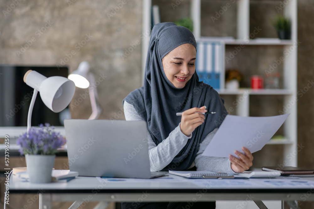 Business Asian muslim woman using calculator and writing make note with calculate doing math finance on an office desk. Woman working at office with laptop and tax, accounting, documents on desk Business Asian muslim woman using calculator and writing make note with calculate doing math finance on an office desk. Woman working at office with laptop and tax, accounting, documents on desk