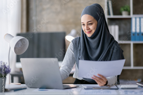 Business Asian muslim woman using calculator and writing make note with calculate doing math finance on an office desk. Woman working at office with laptop and tax, accounting, documents on desk