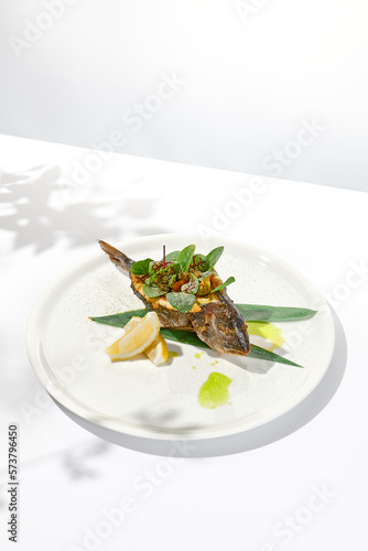 Whole roasted fish with spinach leaves on white plate. Grilled dorado on light background in summer menu. Healthy food - Roasted dorado fish. Concept menu with hard shadows.