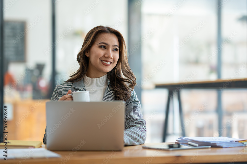 Beautiful asian businesswoman sitting working with laptop computer on table, holding cup of coffee, taking break, relaxing after working on finances. and marketing planning in the office.