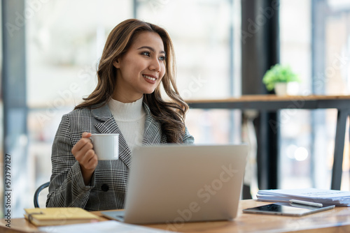 Beautiful asian businesswoman sitting working with laptop computer on table  holding cup of coffee  taking break  relaxing after working on finances. and marketing planning in the office.