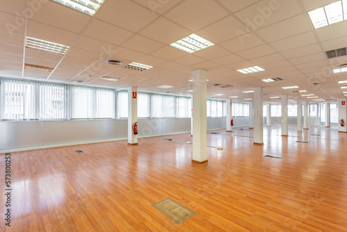 Large empty space with ceiling tiles, fluorescent lights, light brown laminate flooring and white painted columns. Large office space left from a company.