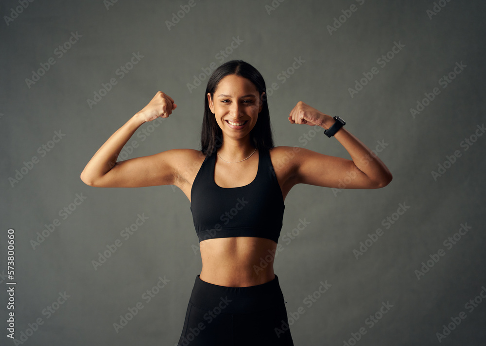 Happy young biracial woman wearing sports bra and fitness tracker flexing biceps in studio Happy young biracial woman wearing sports bra and fitness tracker flexing biceps in studio