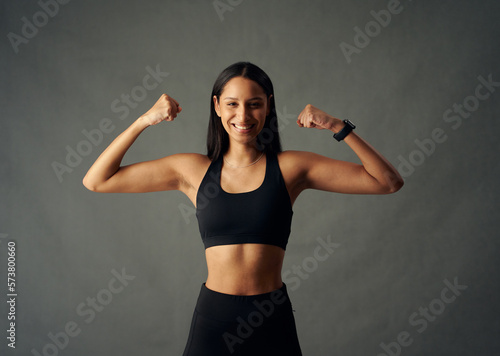 Happy young biracial woman wearing sports bra and fitness tracker flexing biceps in studio