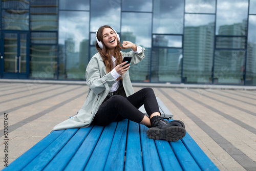 a cheerful young woman listens to music in headphones using a phone while sitting in front of the glass facade of the building on a bench
