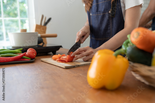 Beautiful Asian woman in kitchen cooking apron preparing various vegetable ingredients and slicing tomatoes in online cooking prep for the health and happiness of our loved ones in their own homes.