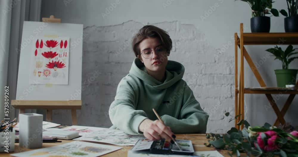 Young stylish woman artist paints pictures on paper with watercolor paints while working in workshop in daylight Young stylish woman artist paints pictures on paper with watercolor paints while working in workshop in daylight