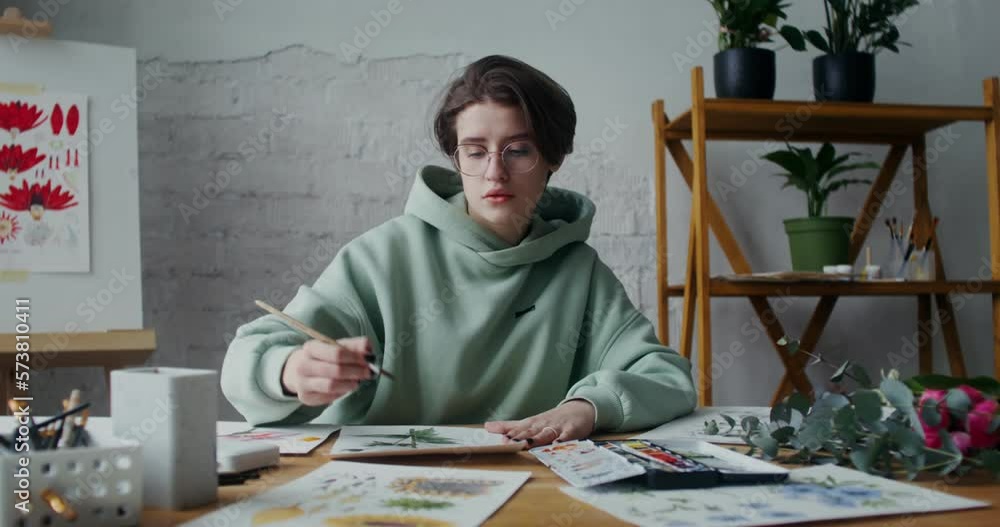 A young woman in glasses draws flowers with watercolors on paper, washing the brush in water from time to time A young woman in glasses draws flowers with watercolors on paper, washing the brush in water from time to time