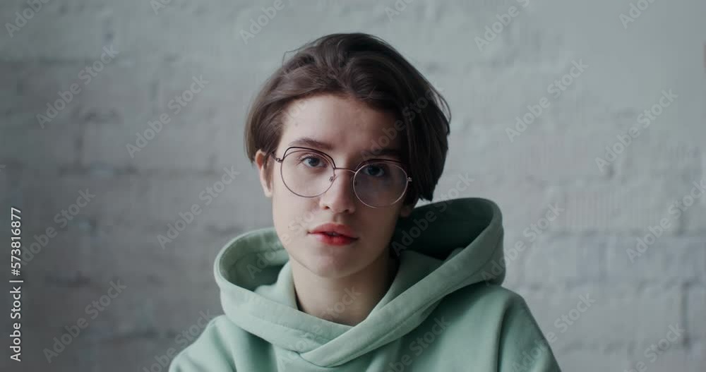 A young woman in glasses and wearing a hoodie looks at the camera with a serious look. Portrait against a brick wall indoors A young woman in glasses and wearing a hoodie looks at the camera with a serious look. Portrait against a brick wall indoors