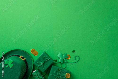 Saint Patrick's Day concept. Top view photo of green leprechaun hat spool of twine present boxes gold coins shamrocks and clover shaped confetti on isolated green background with copyspace