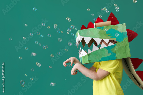 Fotografia Little boy in cardboard dinosaur costume and soap bubbles on green background