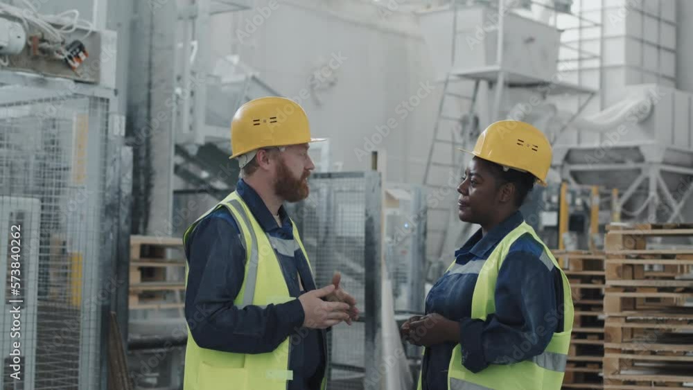Side view of mature Caucasian man and Black woman working together in factory workshop having conversation Side view of mature Caucasian man and Black woman working together in factory workshop having conversation