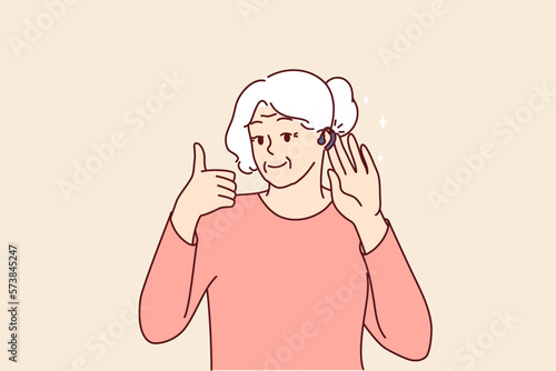 Elderly woman uses hearing aid and shows thumbs up confirming good sound volume thanks to new device. Gray-haired old lady with hearing aid enjoys opportunity to hear others and communicate freely 