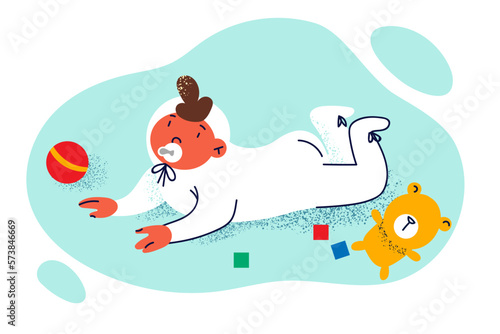 Newborn baby with pacifier in mouth crawls on floor among children toys and dangles legs