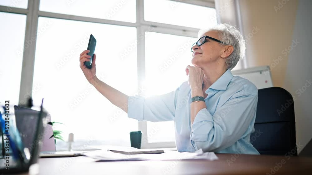 Happy businesswoman wearing eyeglasses taking selfie photo on cellphone and smiling while sitting at desk in office. Adult business lady day off make take selfies on phone. Happy businesswoman wearing eyeglasses taking selfie photo on cellphone and smiling while sitting at desk in office. Adult business lady day off make take selfies on phone.