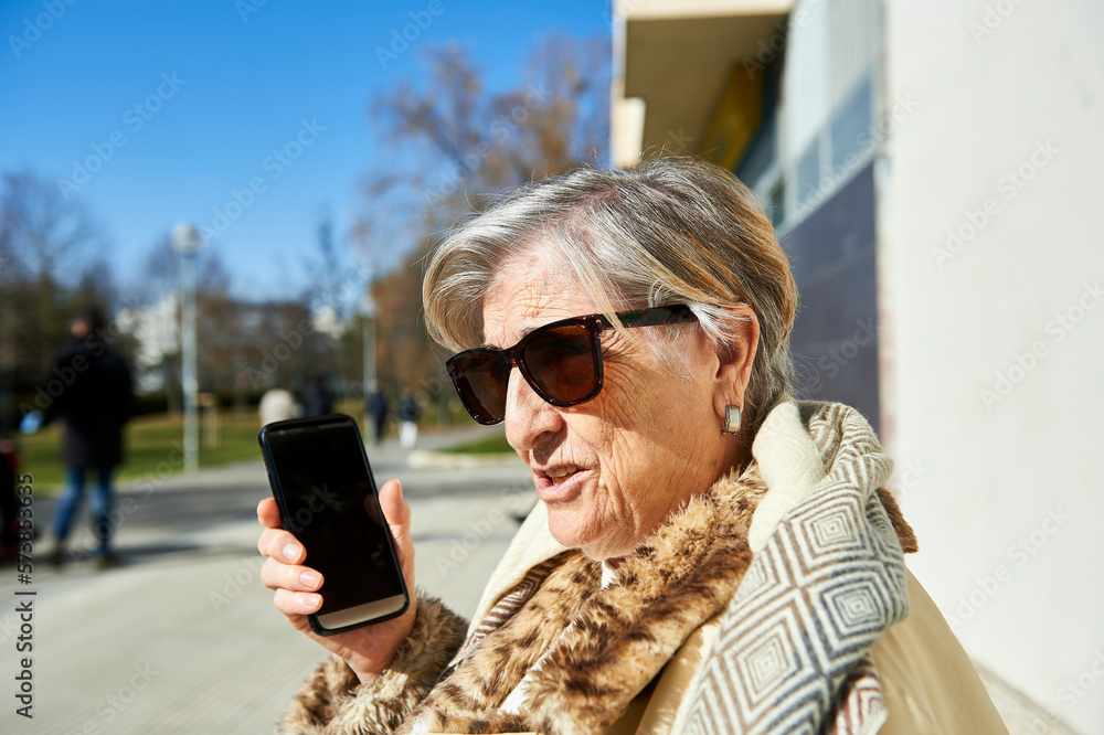 Fototapeta premium an eighty-year-old woman sunbathes in sunglasses sitting on a bench in front of a concrete wall while talking on the cellular phone