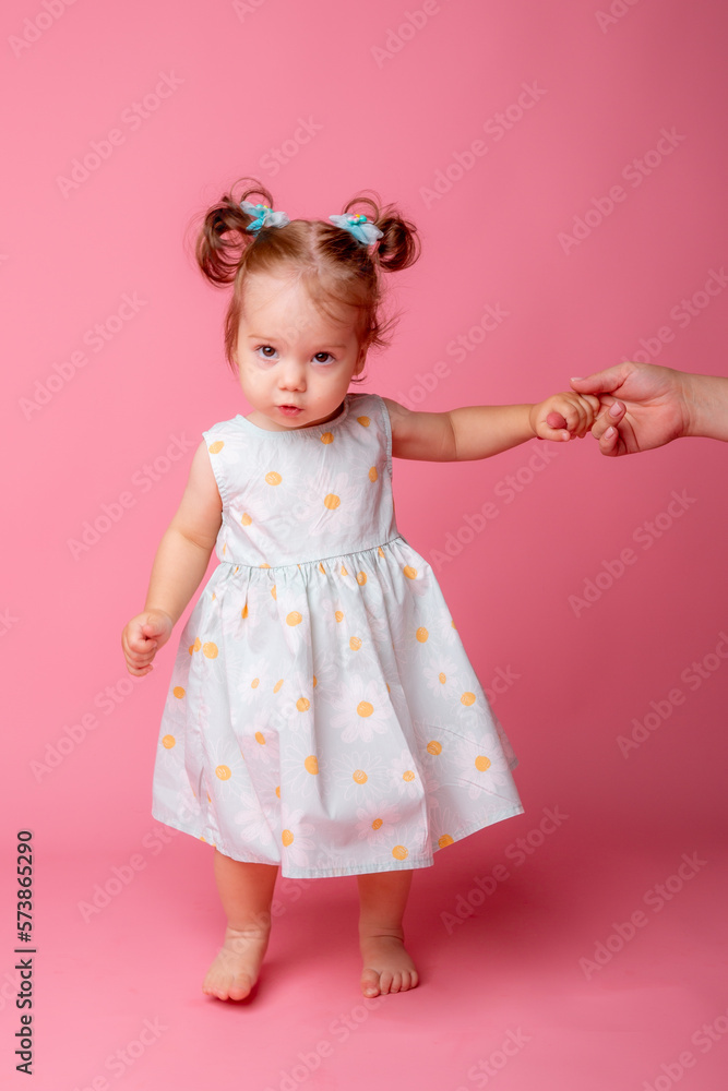 a baby girl stands on a pink background holding her mother's hand