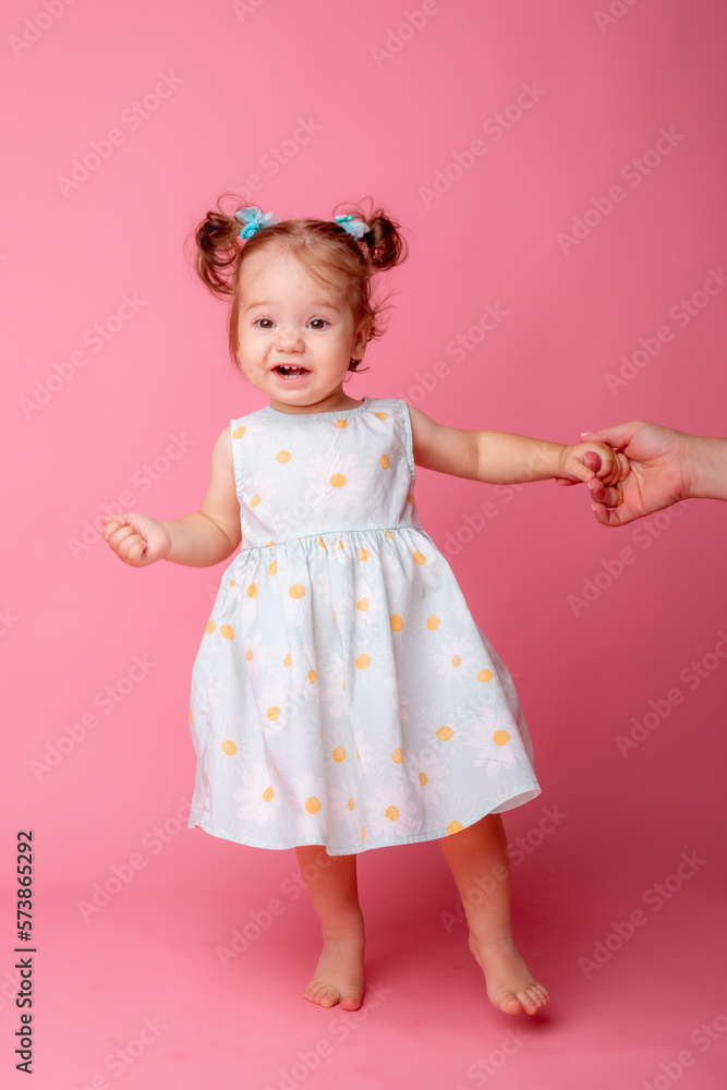 a baby girl stands on a pink background holding her mother's hand