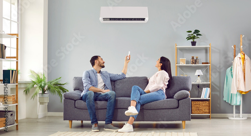 Young happy man and woman turning on air conditioner sitting on sofa at home. Smiling couple of homeowners enjoying cool conditioned air using remote resting on couch together in living room. Banner.
