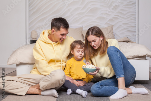 Friendly family, lovely parents with child son sitting by the bed and holding bowl with popcorn