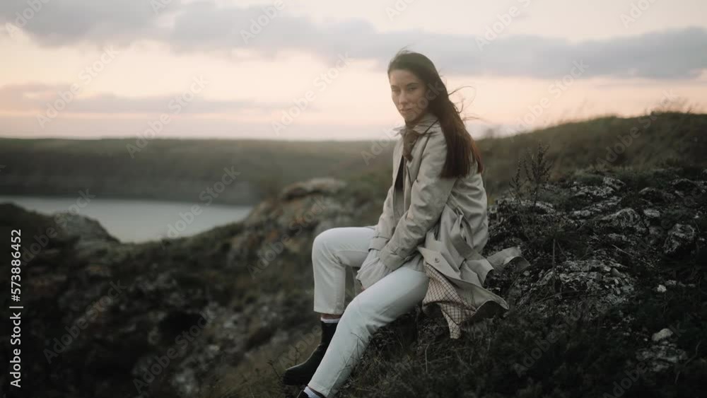 Girl Sitting on a Cliff overlooking the Landscape with her Hair Blowing. Young Woman sitting on a Rock and Looking to the Horizon Girl Sitting on a Cliff overlooking the Landscape with her Hair Blowing. Young Woman sitting on a Rock and Looking to the Horizon