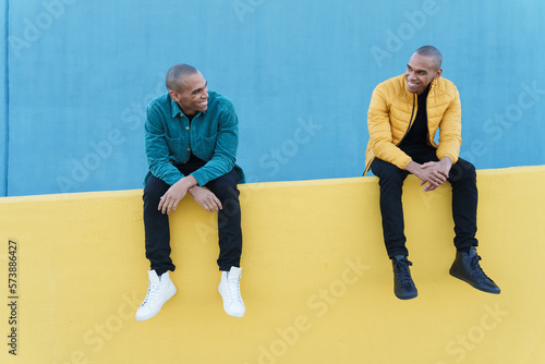 Smiling African American men brothers looking at each other while sitting on yellow concrete fence against blue yellow wall photo