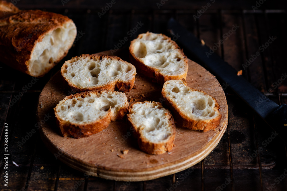 Sliced French baguette on a wooden board on a dark background. Bread with big holes. Preparing for bruschetta. Sliced French baguette on a wooden board on a dark background. Bread with big holes. Preparing for bruschetta.