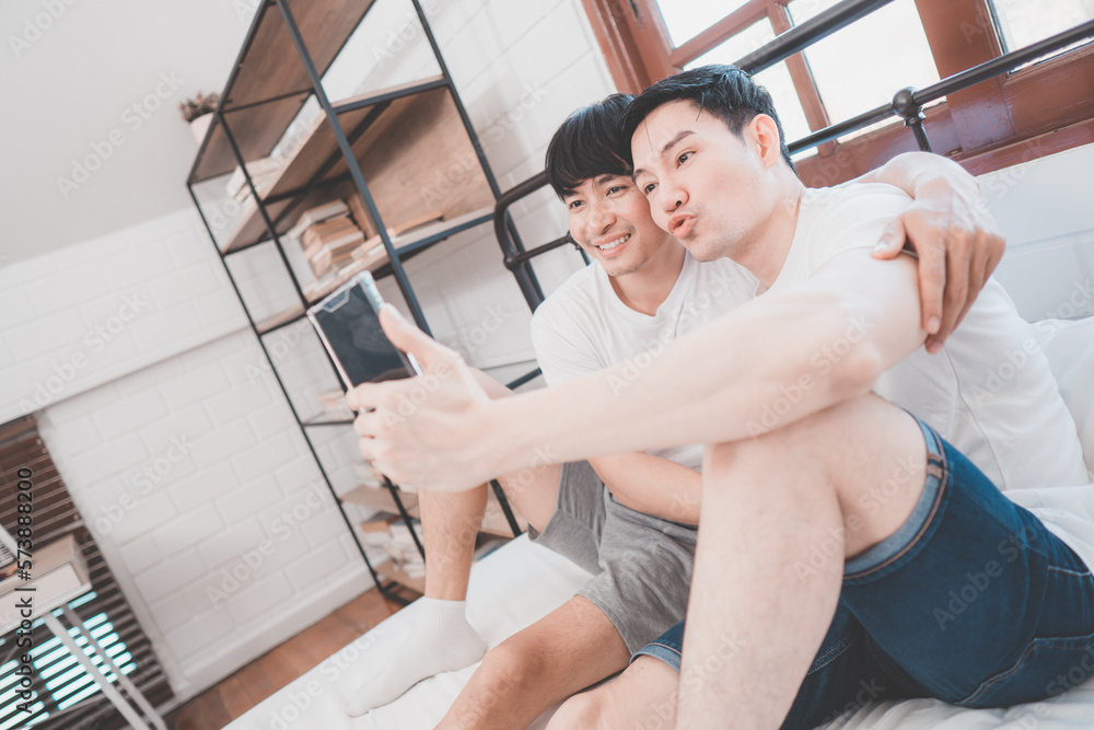 Happy Asian gay couple use mobile phone video call or selfie on bed, LGBTQ concept. Happy gay couple having fun using mobile smartphone in bed. Homosexual love and technology concept