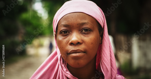 Portrait of Authentic African Woman Standing Under the Rain, Looking at the Camera with a Blurry Greenery Background. Female From a Rural Village in a Headscarf Enjoying the Blessing of Rain photo