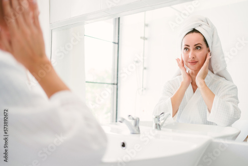 Gorgeous confident adult woman wearing a towel on her head sitting and checking her jaw in front of the mirror in the home bathroom. A pretty attractive woman beauty lifestyle and cosmetic concept.