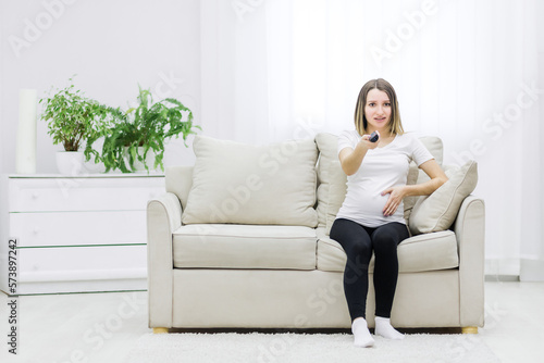 Photo of pregnant young woman with remote control in hand.
